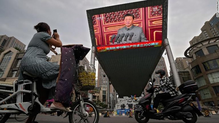 A woman on her electric-powered scooter films a large video screen outside a shopping mall showing Chinese President Xi Jinping speaking